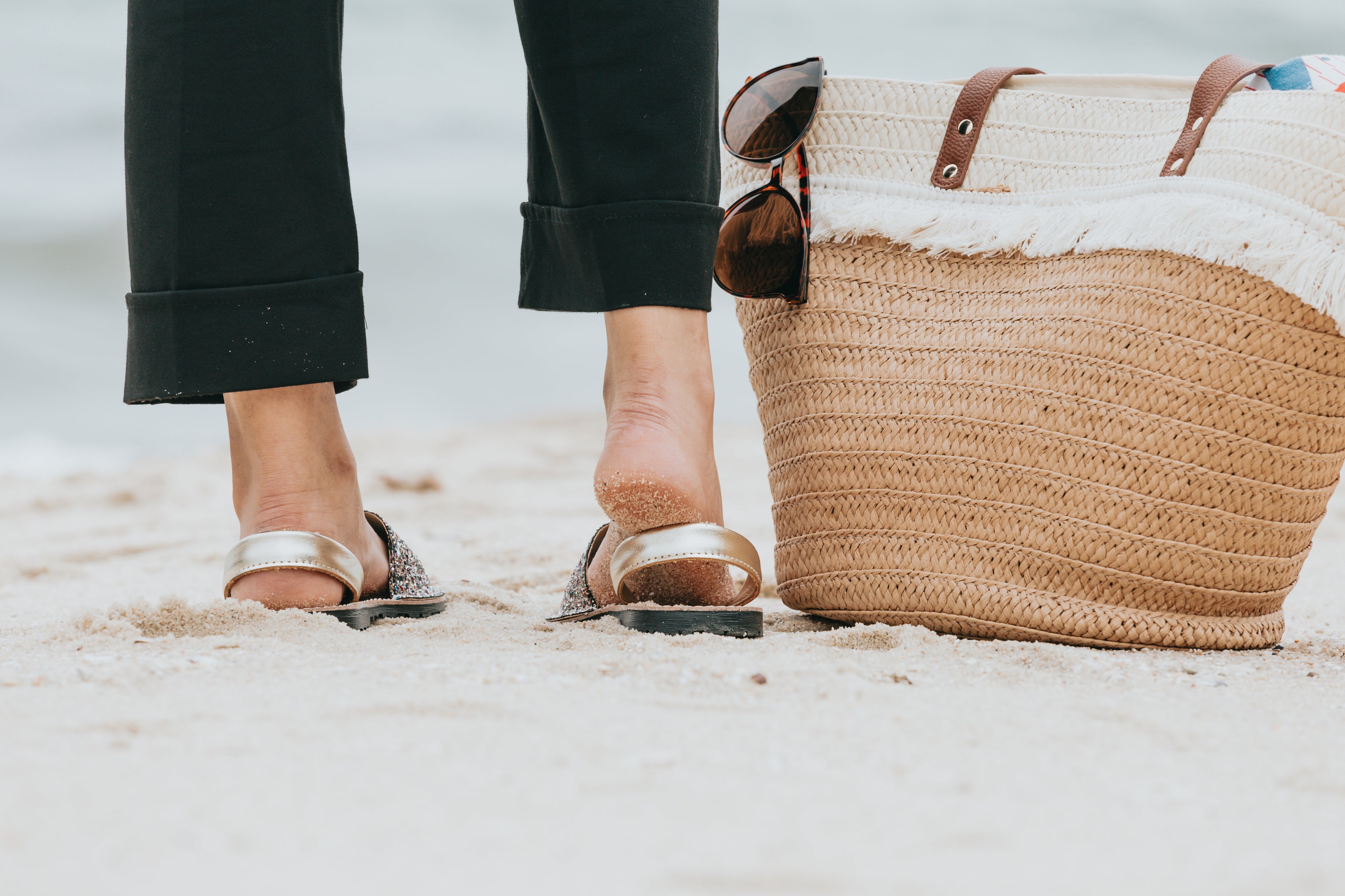 Pieds nus sur une plage de sable à côté d'un cabas en paille tressée Velorie Paris, avec des lunettes de soleil et des sandales dorées